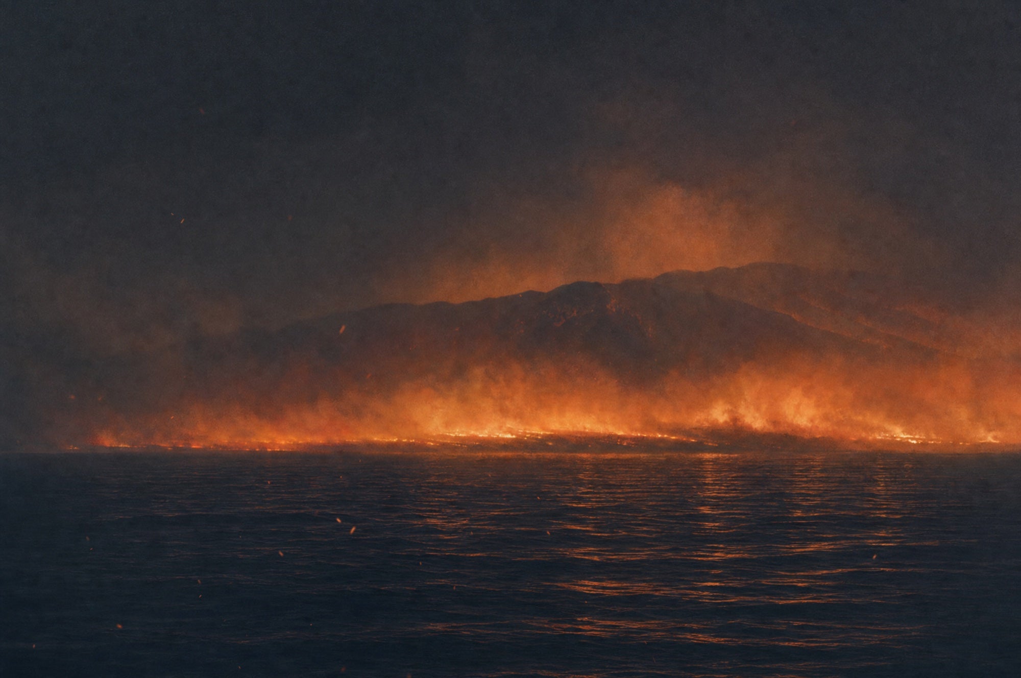A dark ocean at night, and far across it a long low band of fire glow and smoke along a mountainous coast